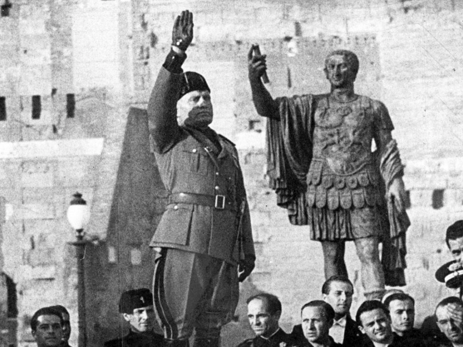Schwarzweißfoto: Mann in Uniform hebt den rechten Arm zum Gruß neben römischer Statue, Zuschauer im Vordergrund.